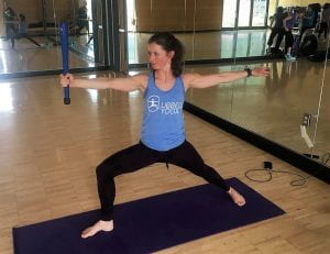 Kate practicing clubbell yoga on a purple mat in a blue shirt
