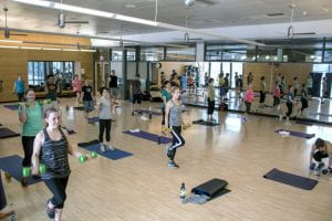 A mirrored room full of fitness folks wearing athletic clothing and holding hand weights while lunging forward.