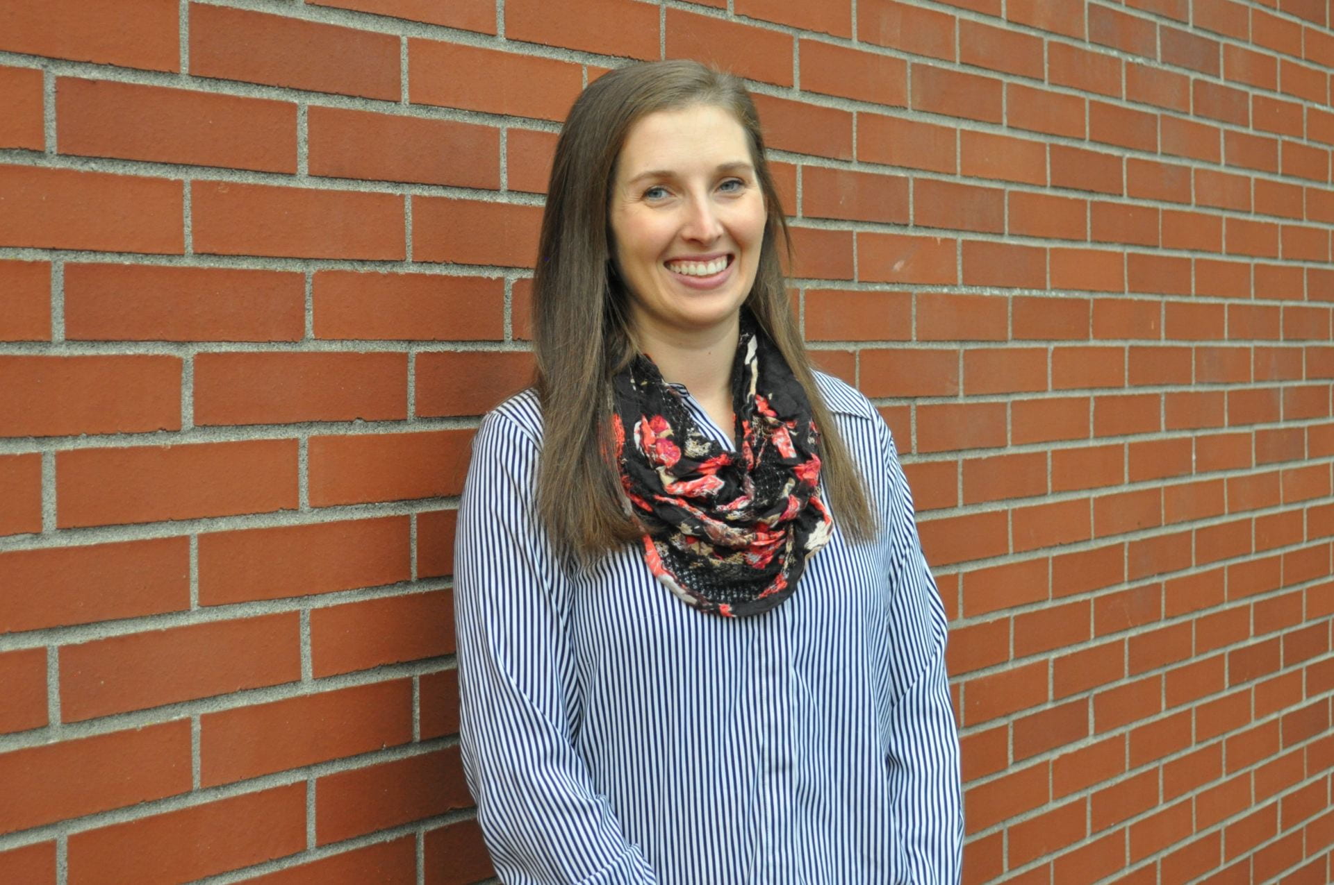 Anna in a grey long sleeve shirt with a patterned scarf standing in front of a brick wall