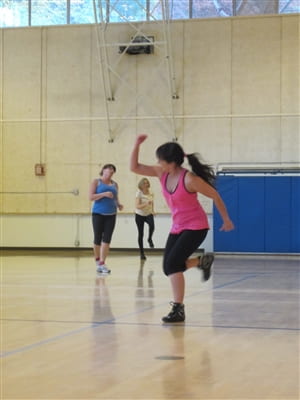 Maria instructing Zumba on a gym court in a pink tank top and black workout pants