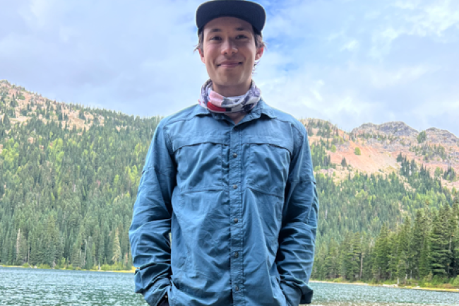 Pierce Bock standing outside in a blue jacket and blue hat in front of a mountain and lake