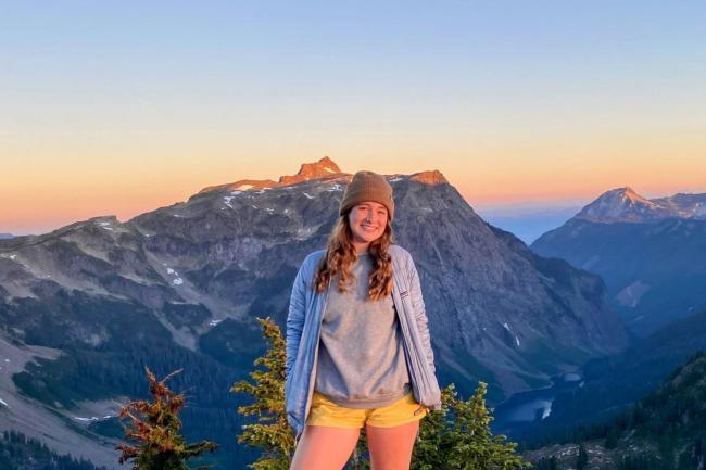 Ruby Gruber standing at the summit of Winchester Mountain in the Mt. Baker-Snoqualmie National Forest with Tomyhoi Peak and Yellow Aster Butte in the background