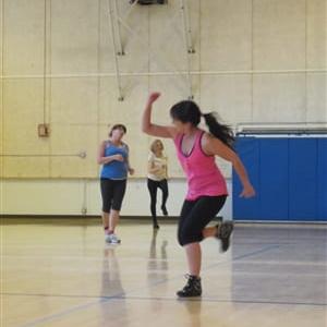 Maria instructing Zumba on a gym court in a pink tank top and black workout pants