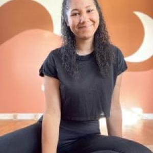 Savanah in a black outfit sitting with her legs bent in front of an orange background